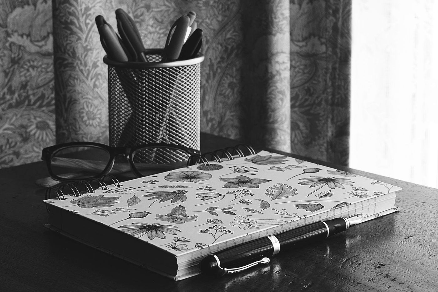 Caderno, óculos e porta-canetas sobre uma mesa, em preto e branco